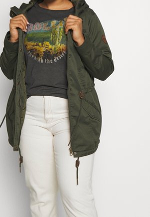 Personne portant une veste à capuche verte par-dessus un t-shirt noir avec un motif de scène du désert et un pantalon blanc, tenant la veste ouverte des deux mains.