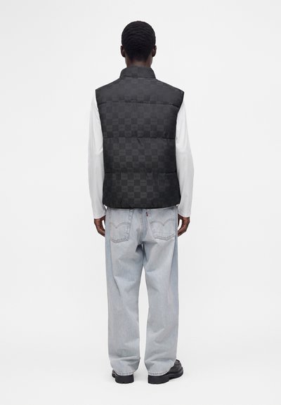 Homme portant un gilet matelassé noir à carreaux par-dessus une chemise blanche à manches longues, un jean bleu clair ample et des chaussures noires, vu de dos sur fond blanc.
