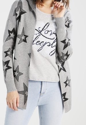 Femme portant un cardigan gris avec des motifs d'étoiles noires sur un t-shirt à cœur pailleté gris clair et un jean bleu clair.