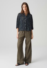 Marineblaues Hemd mit Knöpfen und abstrakten Linien, dreiviertellange Ärmel und lockere Passform, kombiniert mit olivgrünen weit geschnittenen Hosen und schwarzen Sandalen.