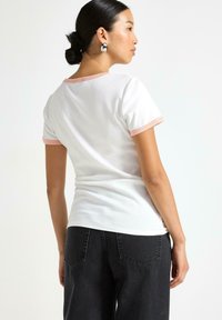 Kvinde med mørkt hår i en knold iført sølvøreringe, en hvid t-shirt med lyserøde kanter og sorte jeans, der står sideværts mod en ensfarvet baggrund.