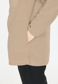 Beige fleecejakke med en struktureret overflade, der har en stor frontlomme. Designet inkluderer en stående krave og lange ærmer.