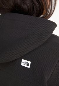 Gros plan sur un sweat à capuche côtelé noir avec un patch logo rectangulaire blanc The North Face sur le haut du dos, juste en dessous de la capuche.