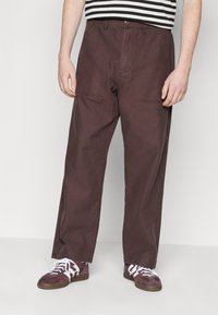Pantalons en toile marron à jambes larges avec deux poches avant, fabriqués à partir d'un tissu durable. Assortis à des chaussures sombres avec des lacets blancs.