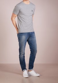 Šedé bavlněné tričko s krátkým rukávem a kulatým výstřihem, s malým černým logem, v kombinaci s modrými džíny a bílými teniskami.