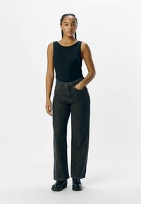 Schwarzes, ärmelloses Top kombiniert mit hoch taillierten, weiten Jeans aus dunkelblauem Denim. Das Model steht vor einem einfarbigen, hellen Hintergrund. Schwarze Knöchelstiefel runden das Outfit ab.