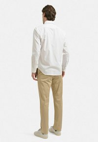 Vit långärmad skjorta med en rundad nederkant, tillsammans med beige byxor och ljusa sneakers, sedd bakifrån.