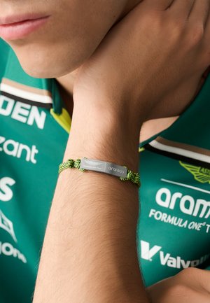 Zöld-fekete fonott karkötő ezüst fém táblával, amelyen a "Formula One Team" és az "aramco" felirat olvasható. Egy csuklón viselve.