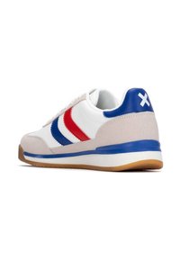 Zapatilla blanca con franjas rojas y azules, acentos de ante, cuello acolchado y suela de goma. Presenta un tirador en el talón azul con detalles en blanco.