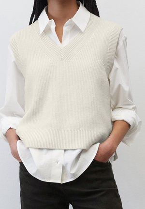 Personne portant une chemise blanche à col sous un pull sans manches en maille crème, les mains dans les poches d’un pantalon noir.