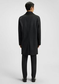 Cappotto in lana a scacchi neri, di lunghezza media, con un solo petto, colletto e tasche laterali. Indossato sopra pantaloni neri slim-fit e stivali.