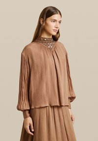 Blusa marrone plissettata con maniche lunghe, caratterizzata da un collo decorato. Tessuto morbido e vestibilità rilassata, abbinata a una gonna di colore simile.