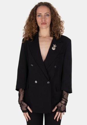 Donna con capelli ricci indossa un blazer nero doppiopetto con una spilla a forma di conchiglia e stella marina e guanti neri in pizzo senza dita.