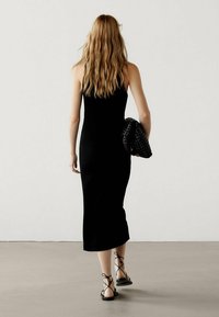 Schwarzes geripptes Midikleid mit einer figurbetonten Silhouette, getragen mit schwarzen Sandalen mit dünnen Riemen und einer geflochtenen schwarzen Handtasche, von hinten betrachtet.