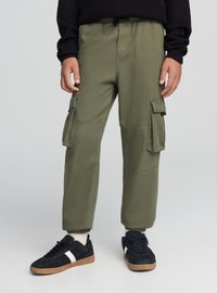 Oliwkowe spodnie bojówki z elastycznymi mankietami, wieloma bocznymi kieszeniami oraz elastycznym paskiem ze sznurkiem, połączone z czarno-białymi tenisówkami.