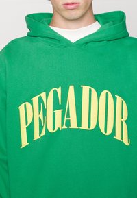 Una persona che indossa una felpa verde brillante con testo giallo che dice "PEGADOR" si trova contro uno sfondo semplice, concentrandosi sull'abbigliamento.