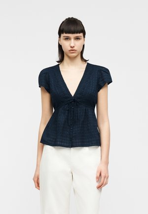 Femeie cu păr drept și închis la culoare, purtând o bluză bleumarin cu decolteu în V și nasturi, precum și pantaloni albi cu croială largă, pe un fundal simplu, deschis la culoare.