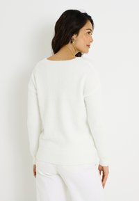 Pull blanc et duveteux avec une coupe décontractée, col rond et manches longues, associé à un pantalon blanc. Texture douce, design minimaliste.