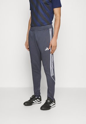 Homme portant un pantalon de survêtement Adidas bleu marine avec des bandes blanches et des chaussures de sport Adidas noires, debout devant un fond blanc.