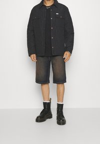 Quilted black jacket with snap buttons, two chest pockets, black t-shirt, dark denim shorts, black lace-up boots with prominent soles.