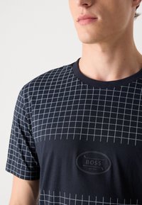 Tamsiai mėlyni trumpomis rankovėmis marškiniai su balta tinklo raštu ir iškilnia juoda logotipo etikete, kurioje antkyrašta "BOSS".