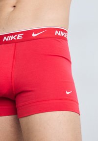 Rote Nike-Boxershorts mit einem weißen elastischen Bund, auf dem das "NIKE"-Logo prangt. Glatte Textur, figurbetonter Schnitt und Logo-Details auf den Shorts.