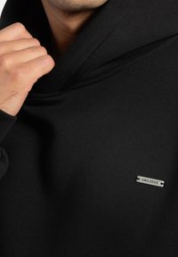 Mann, der die Kapuze eines schwarzen Sweatshirts anpasst, auf dem sich ein kleines rechteckiges Metallschild mit der Aufschrift "SMILODOX" auf der Brust befindet.