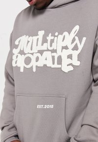 Šedá mikina s texturovaným bílým nápisem "Multiply" na hrudi a "EST. 2015" vytištěným v dolní části kapsy. Měkký materiál.