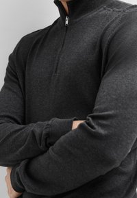 Tmavě šedá mikina na zip s vysokým límcem. Materiál vypadá měkký a pletený s žebrovanými manžetami a detaily límce. Minimalistický design.