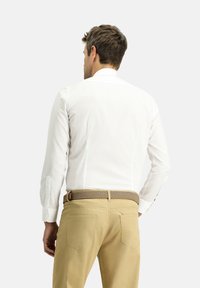 Camicia bianca a maniche lunghe con colletto a punta, dotata di polsini con bottoni. Abbinata a pantaloni beige e una cintura intrecciata marrone. Vista posteriore.