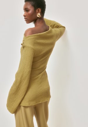 Sieviete ar īsiem matiem valkā teksturētu mustarda krāsas džemperi ar atsegtiem pleciem un bēšas krāsas bikses, pagriežas sāniski, ar roku aiz galvas, redzamas zelta abstraktas auskaras.