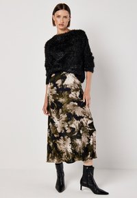 Schwarzer flauschiger Pullover mit kurzen Ärmeln, kombiniert mit einem langen, floralen Rock in Erdtönen. Das Model trägt schwarze Stiefeletten mit Reißverschluss.