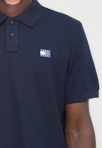 Nehéz anyagból készült tengerészkék polo ing. Három gombos nyakkivágása van, és a mellkasán egy kis téglalap alakú kék-fehér "TOMMY JEANS" logó található.