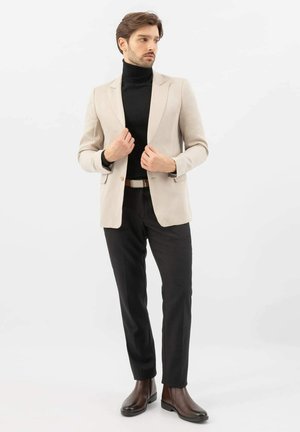 Blazer beige sur un col roulé noir, associé à un pantalon noir et des bottines en cuir marron. Tissu lisse, coupe ajustée, minimalisme des détails.