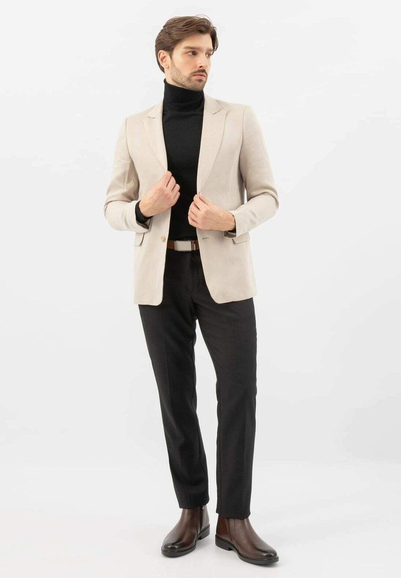 Blazer beige sopra un dolcevita nero, abbinato a pantaloni neri e stivaletti alla caviglia in pelle marrone. Tessuto liscio, vestibilità su misura, dettagli minimal.