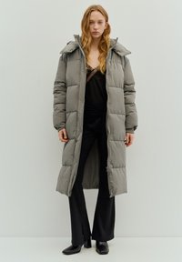 Lange grijze donzen jas met doorgestikt design, vaste capuchon en rits over de volle lengte. Gedragen over een zwart topje en aansluitende zwarte broek.