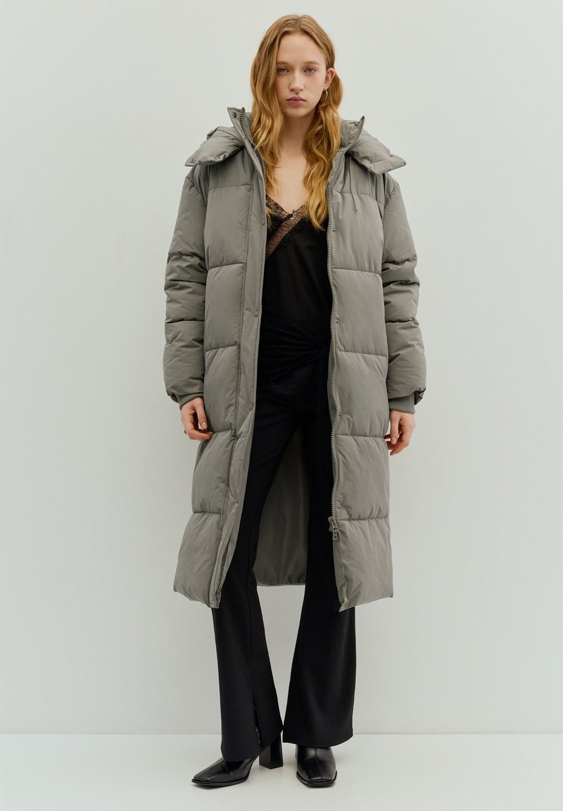 Lange grijze donzen jas met doorgestikt design, vaste capuchon en rits over de volle lengte. Gedragen over een zwart topje en aansluitende zwarte broek.