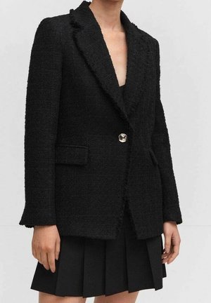 Kvinde iført en tekstureret sort blazer med en enkelt metallisk knap over et sort plisseret skørt, stående mod en ensartet lyse baggrund.