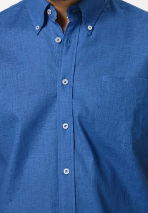 Camisa de botón azul hecha de tejido texturizado, con un pequeño bolsillo en el pecho y botones de color azul claro. El cuello está doblado hacia abajo.