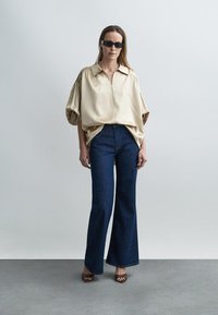 Blusa bege de cetim com gola e mangas bufantes, combinada com jeans azul escuro flare e sandálias castanhas de salto.