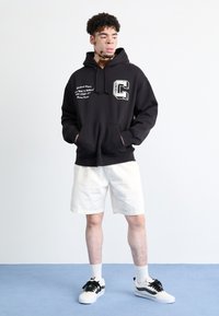 Felpa nera con cappuccio e tasca frontale, lettering bianco e logo "C". Abbinata a pantaloni corti bianchi e scarpe nere e bianche.