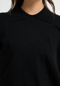 Schwarzes Strickoberteil mit einem kragenförmigen Ausschnitt und schwarzen Perlendetails entlang der Kragenränder. Glatte Textur, taillierte Passform.