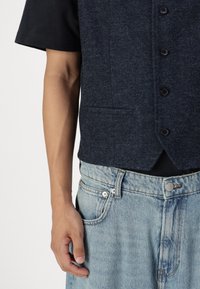 Marine blå vest med tekstur, seks knapper og lommer foran, båret over en svart skjorte, sammen med falmede lyseblå denimjeans.