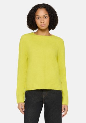 Frau mit lockigem Haar, die einen leuchtend gelben Pullover mit langen Ärmeln und schwarze Jeans trägt, vor einem einfarbigen hellen Hintergrund stehend.