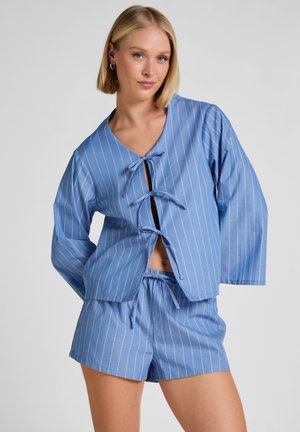 Vrouw draagt een blauw pyjamapak met krijtstrepen, bestaande uit een longsleeve top met drie strikken en bijpassende korte broek, staand tegen een effen achtergrond.