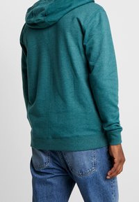 Felpa con cappuccio verde teal, maniche lunghe, polsini e orlo a coste. Tessuto morbido, vestibilità casual e design leggermente oversize.