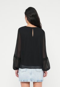 Femme aux longs cheveux foncés porte un chemisier noir transparent à manches longues avec un dos échancré et une jupe en jean bleu clair, debout de dos.
