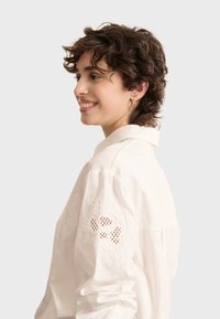 Camicia bianca di cotone con design floreale ricamato sulle maniche. Presenta un colletto classico e una vestibilità rilassata, con una texture morbida e liscia.