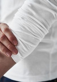 Ilga rankovė balta medvilninė marškiniai su susiraukšlėjusia medžiaga rankovėje, rodo ranką, ilsinčiąsi ant rankos, akcentuojančią lygią tekstūrą ir prigludimą.