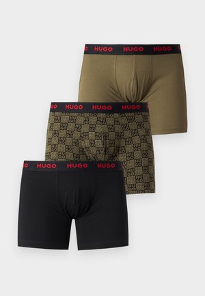 Skjorteløs mand stående barfodet iført sorte boxerbriefs med orange "HUGO" logo mønster og taljebånd på en ensfarvet hvid baggrund.